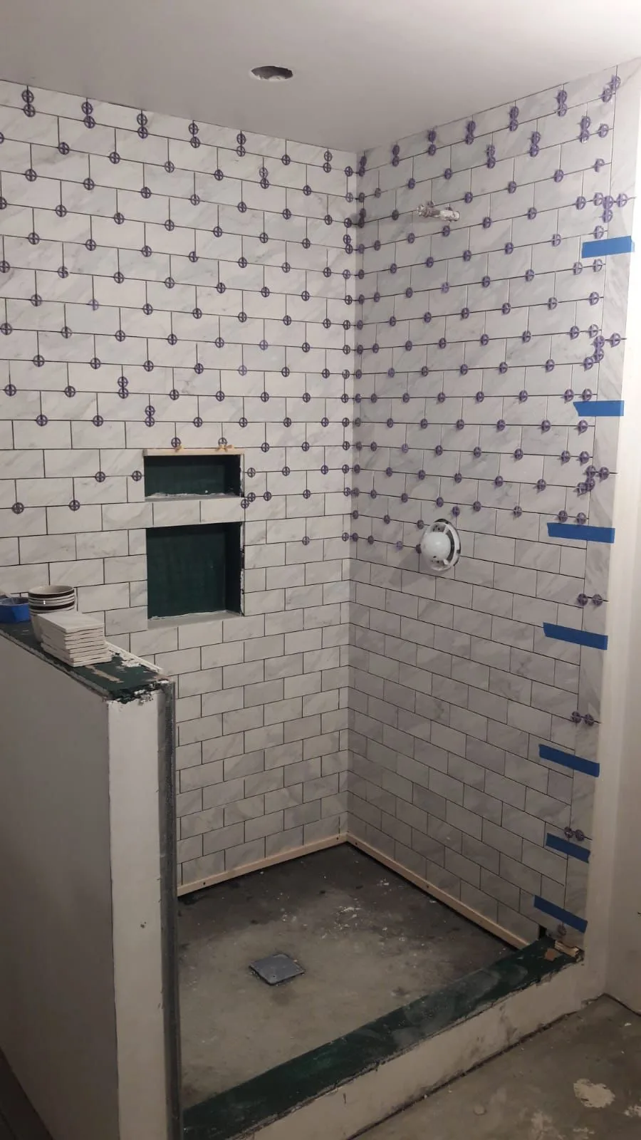 Shower tile installation progress showing marble look subway tiles and leveling spacers around two wall niches.